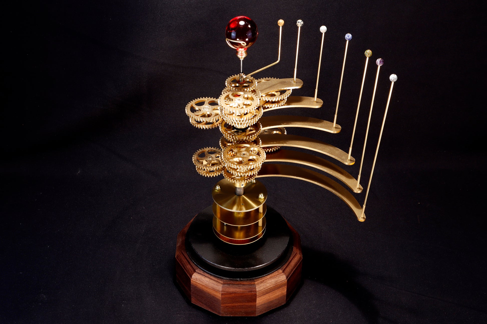 An Orrery model depicting the Trappist-1 system in the constellation Aquarius. A sphere of red glass representing the dwarf star, and semi-precious stones depicting the Trappist-1 planets. Solid brass mechanical gears. The orrery is mounted on a hardwood wooden base.