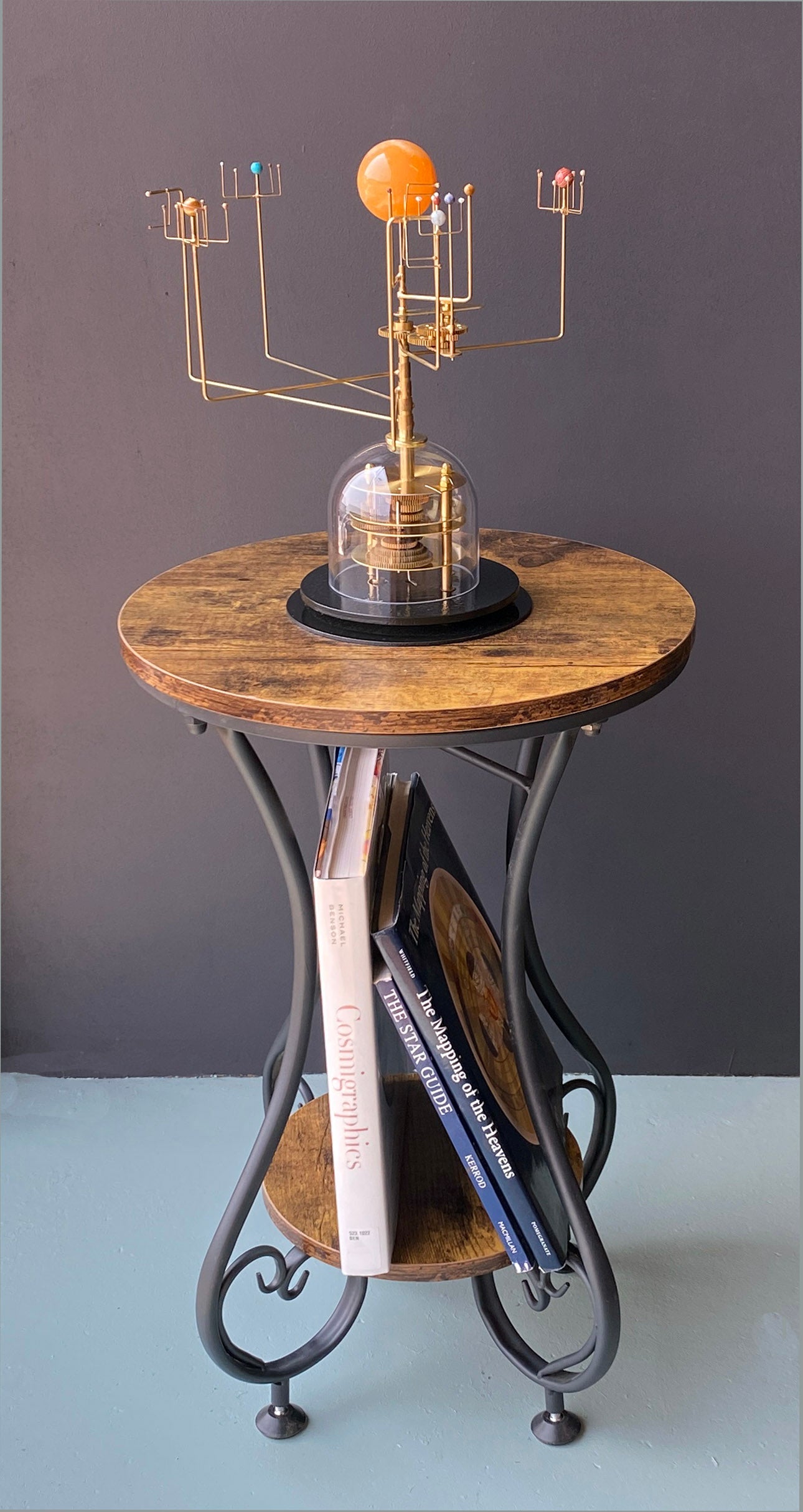 A brass gear mechanism Orrery with semi-precious stone planets mounted on a side table with a black iron frame and a rustic brown tabletop. Books are shown and the smaller table shelf.