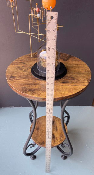A brass gear mechanism Orrery with semi-precious stone planets mounted on a side table with a black iron frame and a rustic brown tabletop. A large yard stick is shown against the table to show size and height.