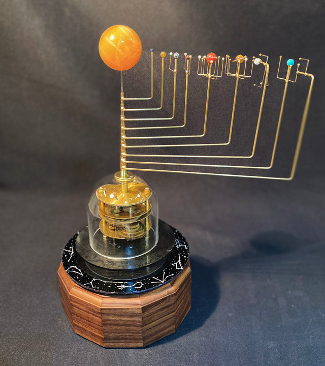 A solar powered orrery model featuring a glass dome, brass gears, semi-precious stone planets in orbit around an orange calcite sun, mounted on a hardwood base.